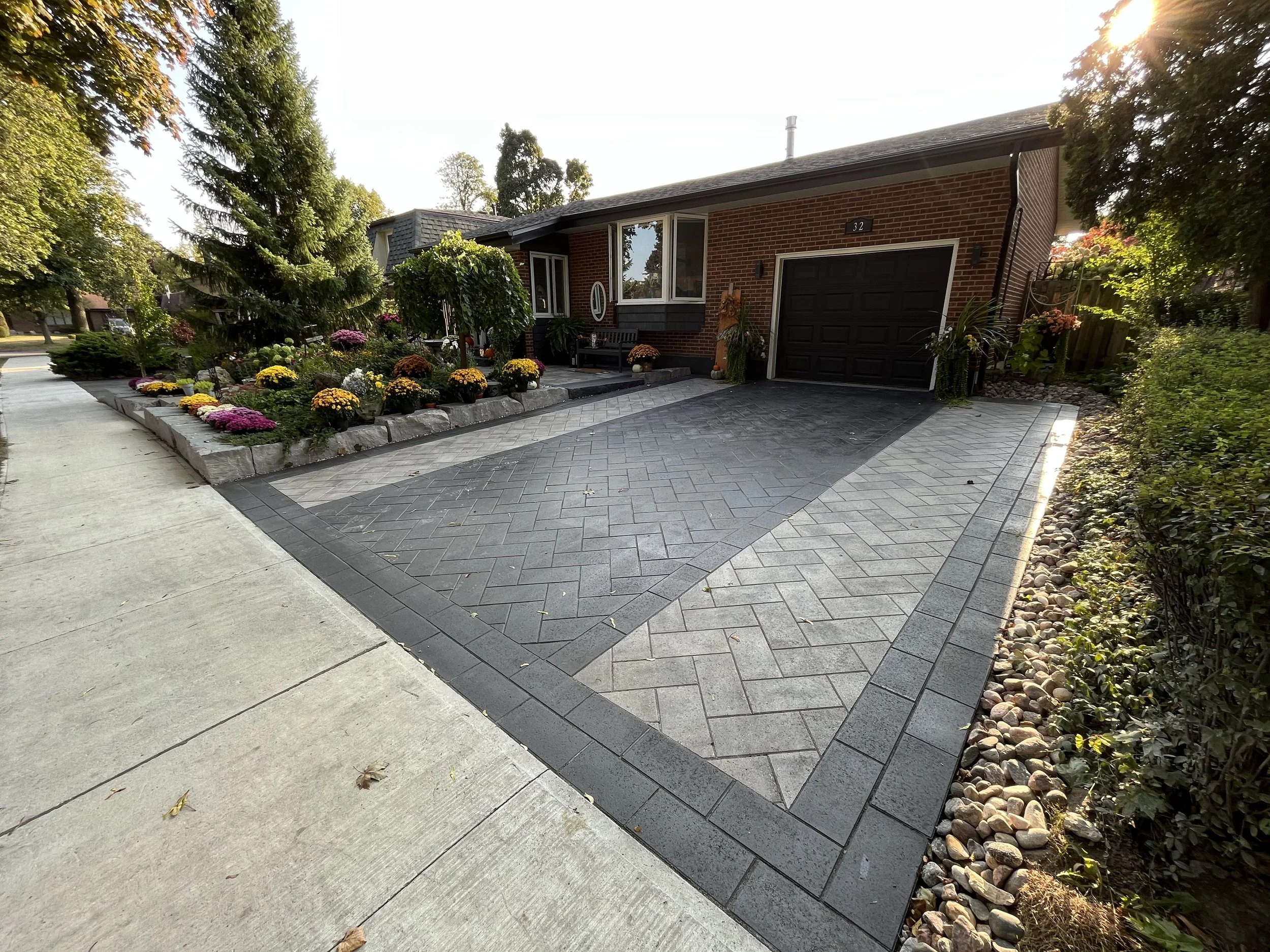 Front yard of a brick house with a black garage door, paved driveway, flower bed with colorful chrysanthemums, trees, and a concrete sidewalk.
