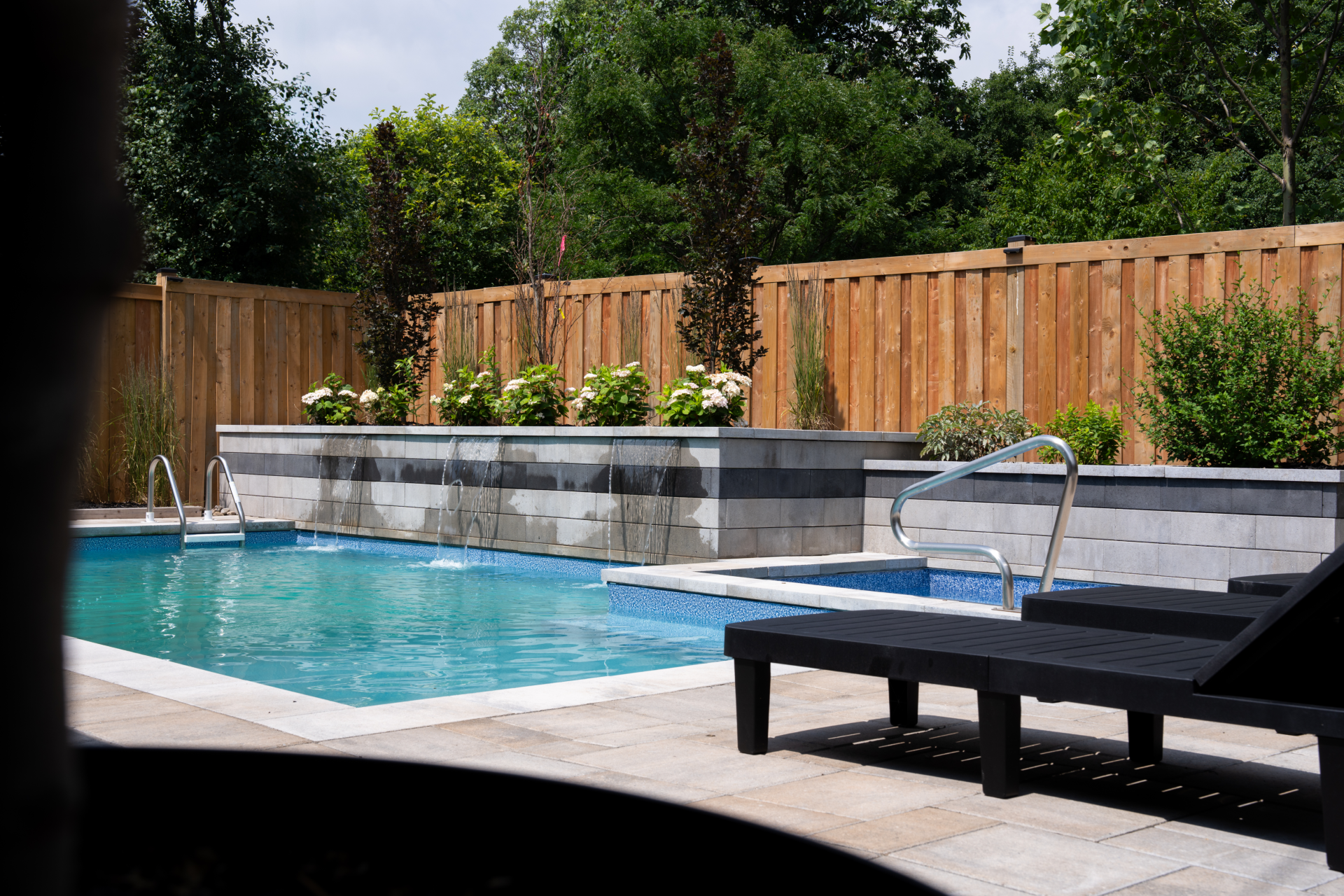 A backyard swimming pool with a water feature, surrounded by a wooden fence and lush greenery, with two black lounge chairs on a patio.