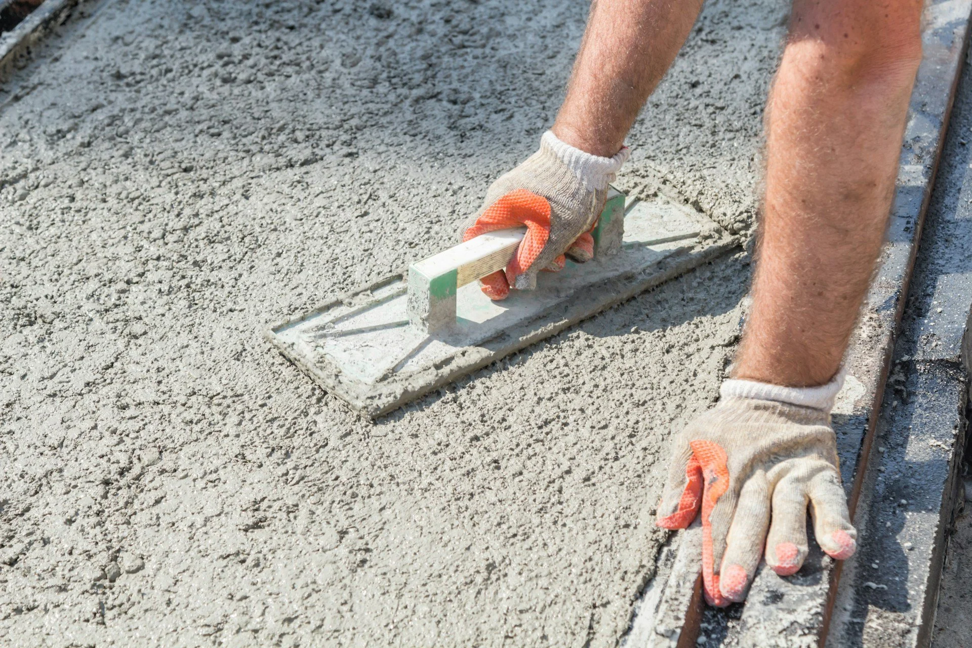 Person smoothing fresh concrete with a float during construction, wearing gloves and kneeling on the ground.