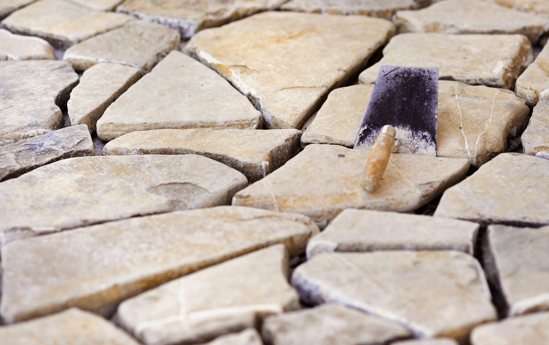 Close-up of beige natural stone pavers with a grout chisel and hammer on the stone surface, used for stone paving or tile installation.