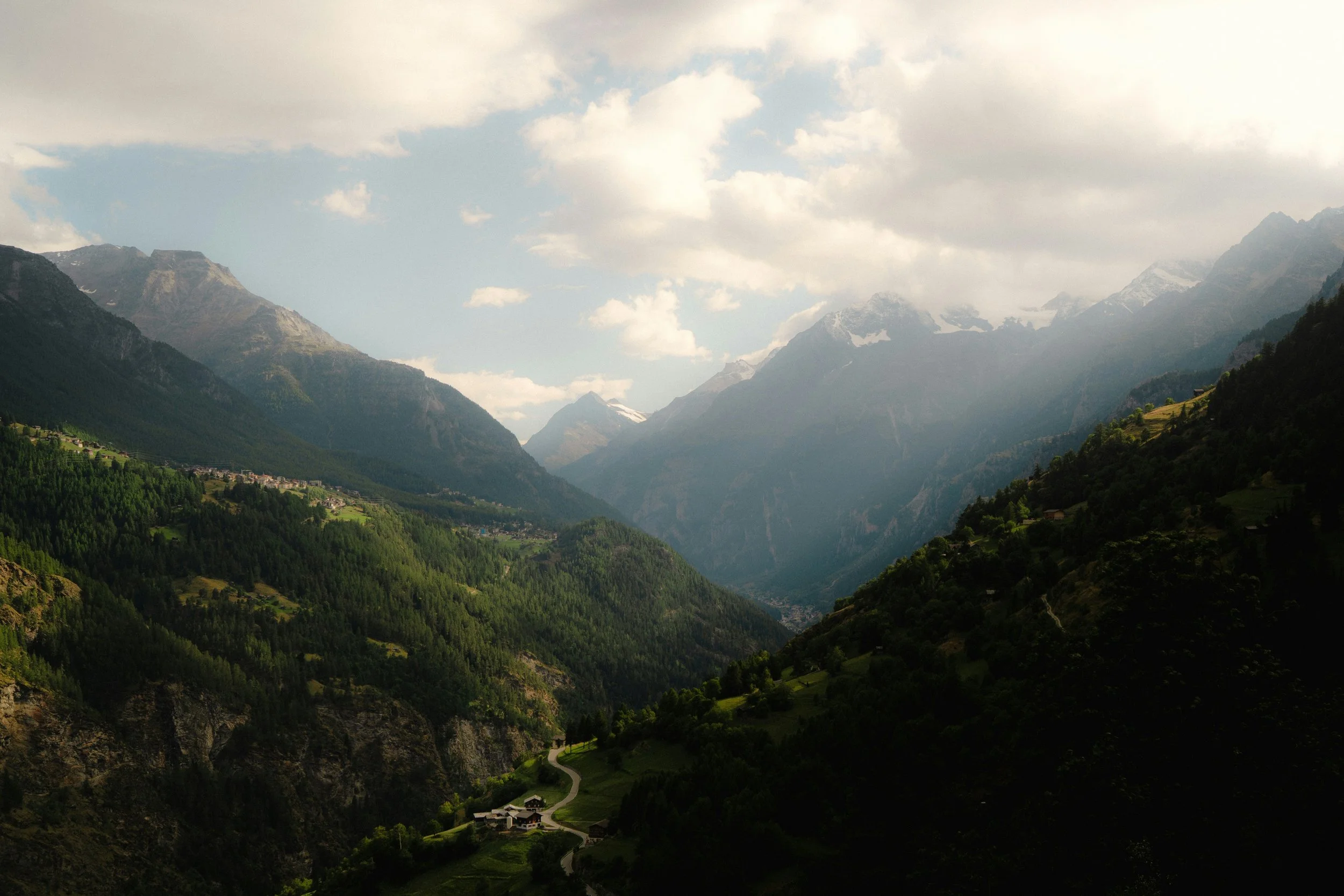 Paysage de montagnes avec vallées verdoyantes, un ciel partiellement nuageux et une petite ferme dans la vallée.
