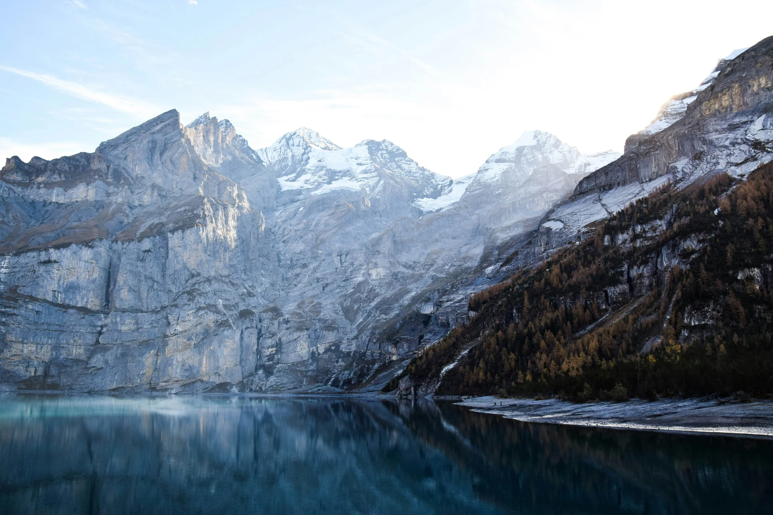 Montagnes enneigées au bord d'un lac calme, forêts sur les pentes et ciel clair