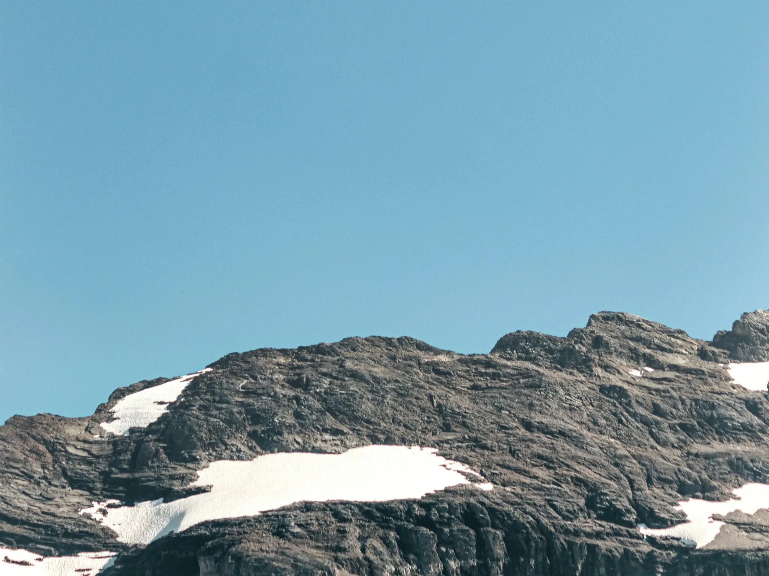 Photo d'une montagne rocheuse avec des zones de neige, sous un ciel bleu clair.