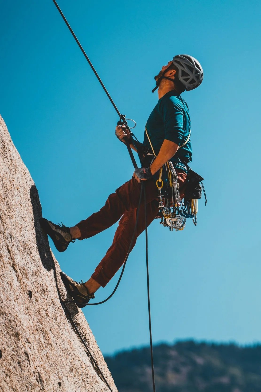 Un grimpeur en activité, équipé d'un casque et d'un harnais, grimpe une paroi rocheuse avec du matériel d'escalade, sous un ciel clair.