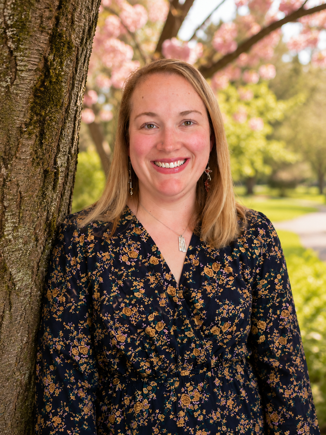 A smiling woman with light brown hair is standing next to a tree in a park with pink flowering trees in the background.
