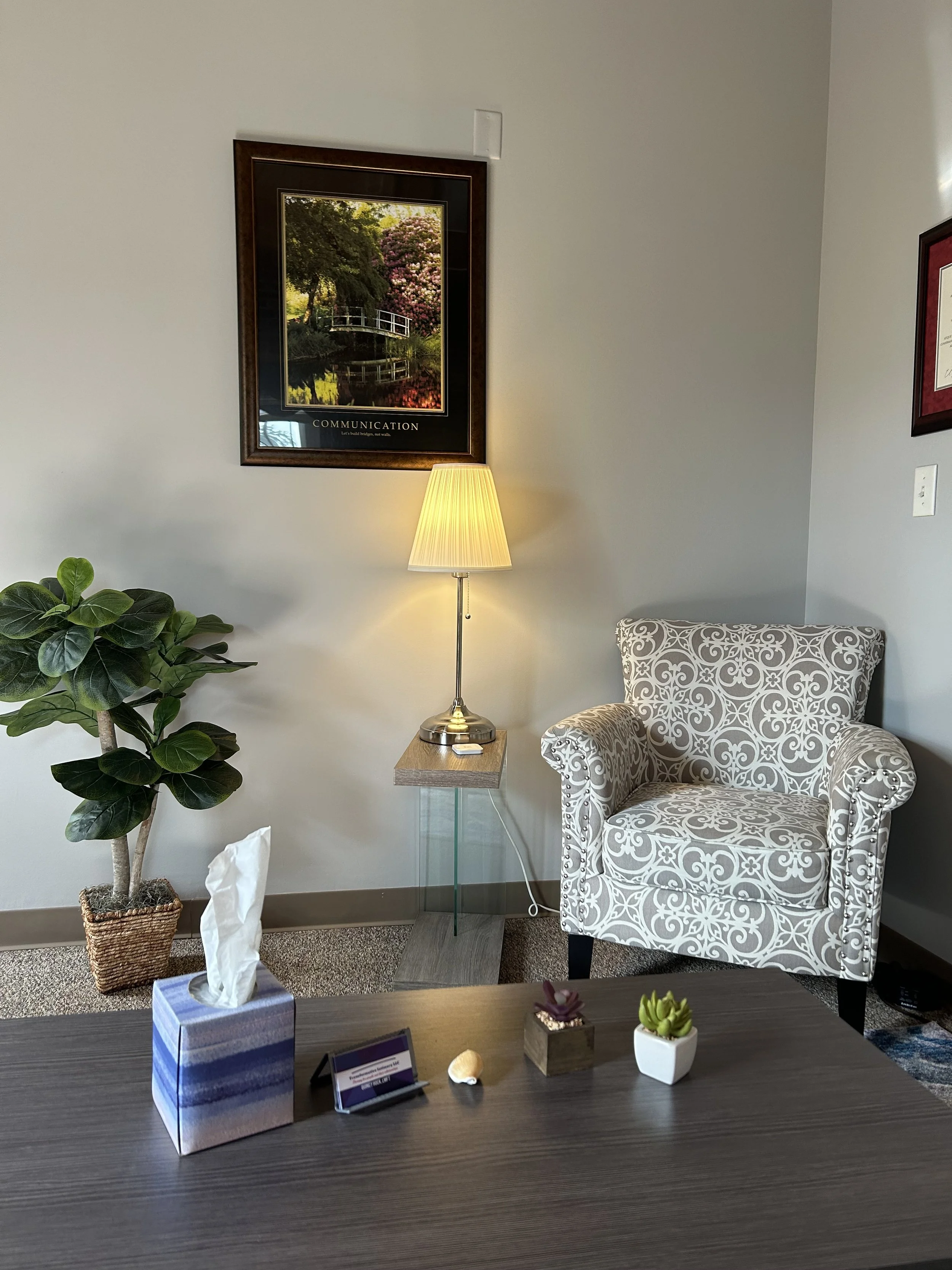 An office with a patterned armchair, a side table with a lamp, plants, and framed artwork on the walls.