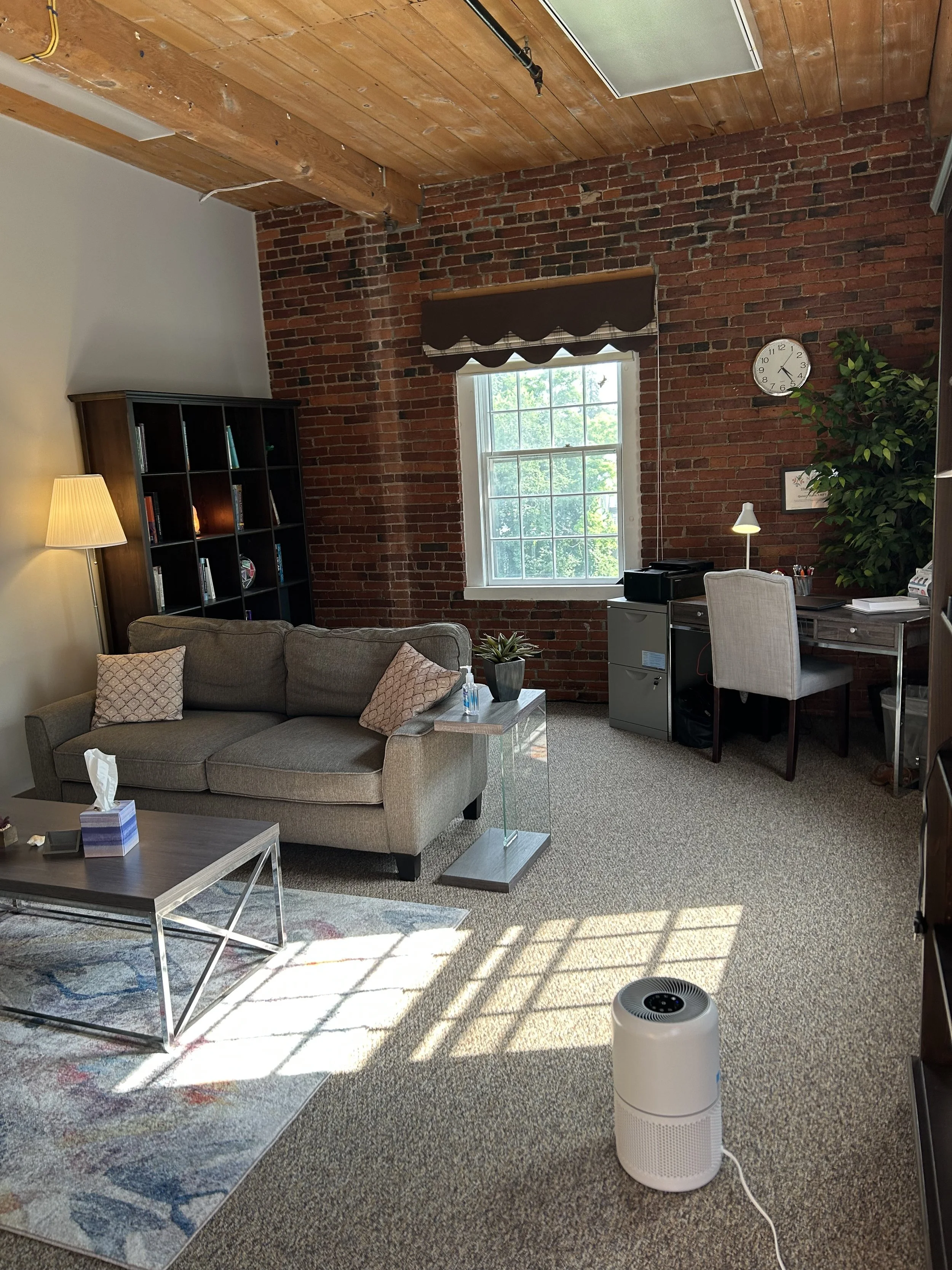 A cozy living room featuring a gray sofa with patterned pillows, a coffee table, a black bookshelf, a window with a brown valance, a desk with a chair, a clock on an exposed brick wall, a tall green plant, a floor lamp, a desk lamp, and a white air purifier on a carpeted floor with sunlight streaming in.