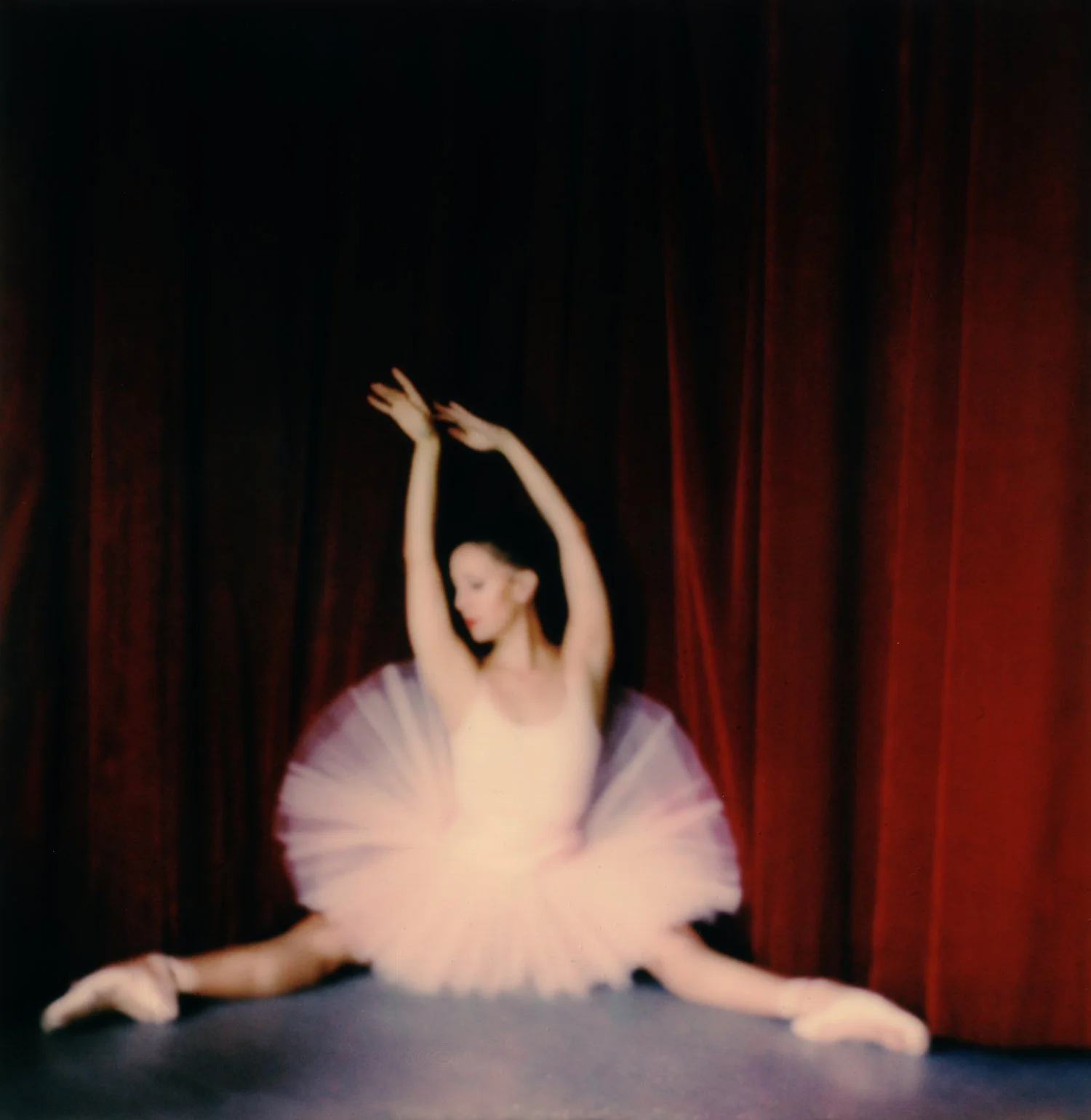 A ballet dancer in a pink tutu performing a split on stage with a red curtain in the background.