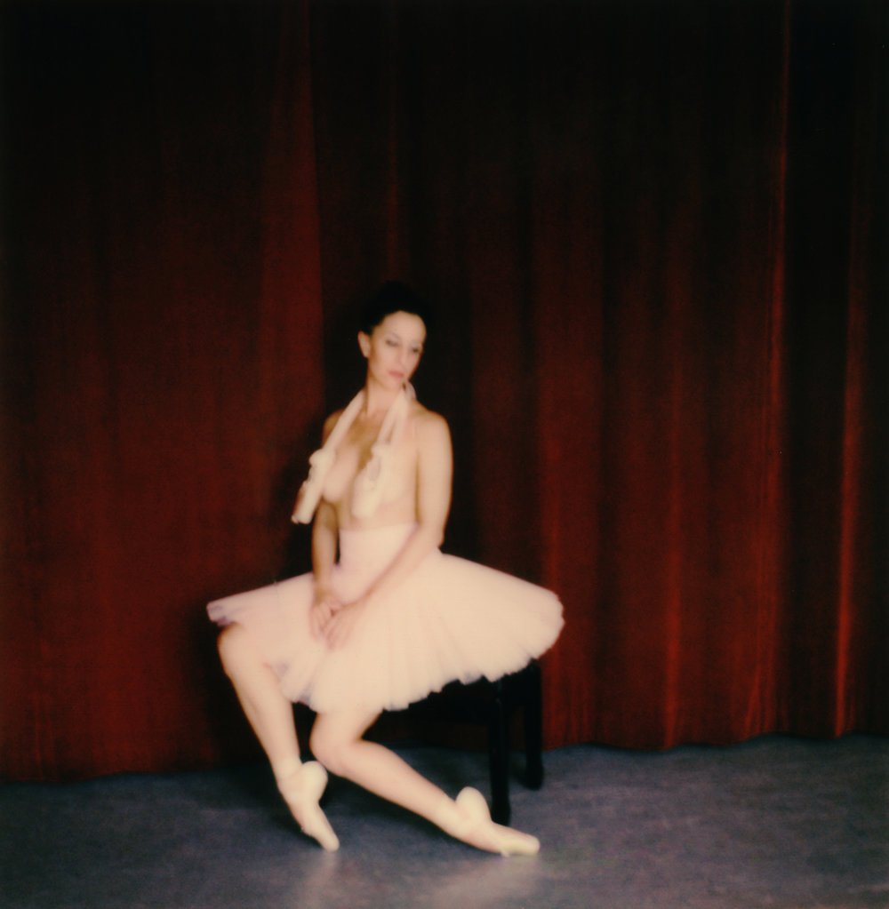A ballerina sitting on a black chair in front of red curtains, wearing a light pink tutu and ballet slippers.