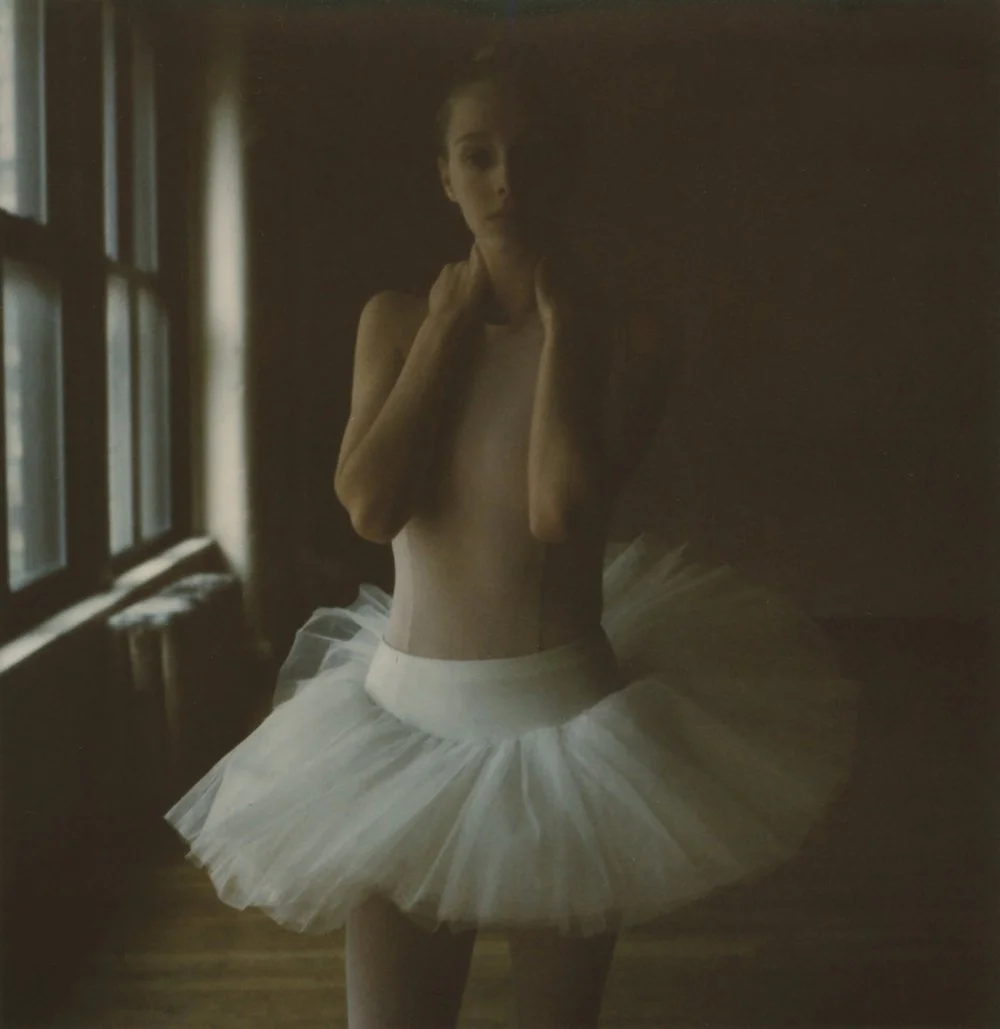 A woman wearing a white tutu skirt standing near a window in a dimly lit room.