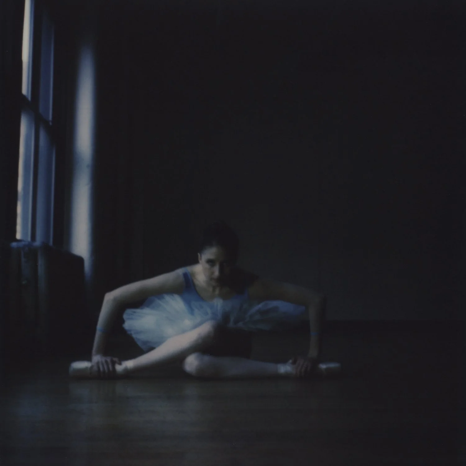 A ballerina in a blue tutu stretching on a dark dance studio floor with large windows letting in natural light.