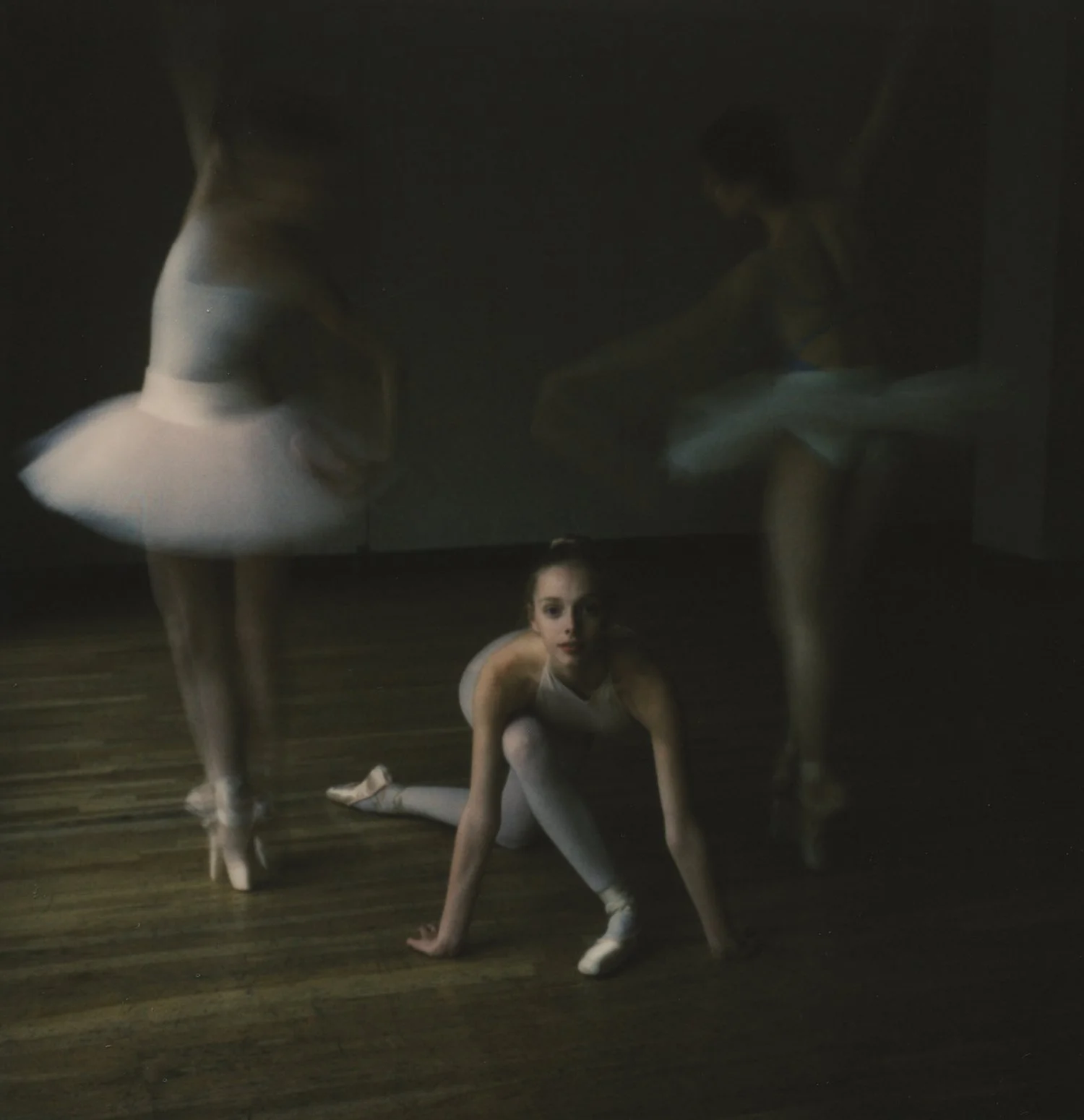 A ballet dancer in a white tutu crouching on the wooden floor, with two other ballerinas reflected in a mirror behind her, dressed in similar outfits, practicing ballet moves.