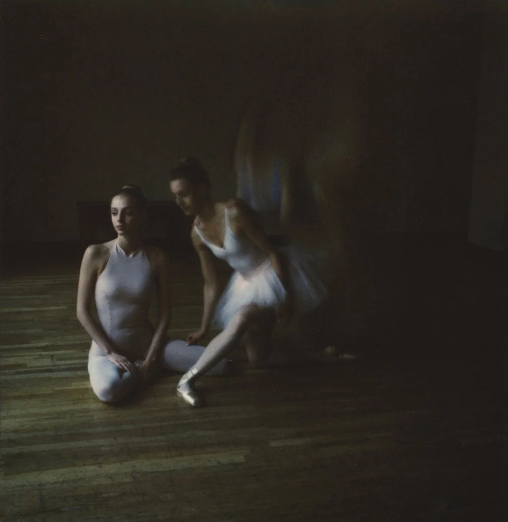 Two ballet dancers in white leotards, one kneeling and the other standing, in a dimly lit studio with wooden floor.