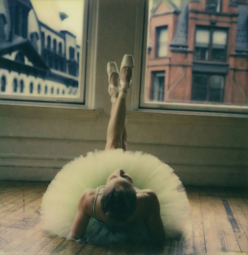 A ballerina in a tutu and ballet slippers lying on her back on a wooden floor, stretching her leg upward, in front of large windows showing city buildings.