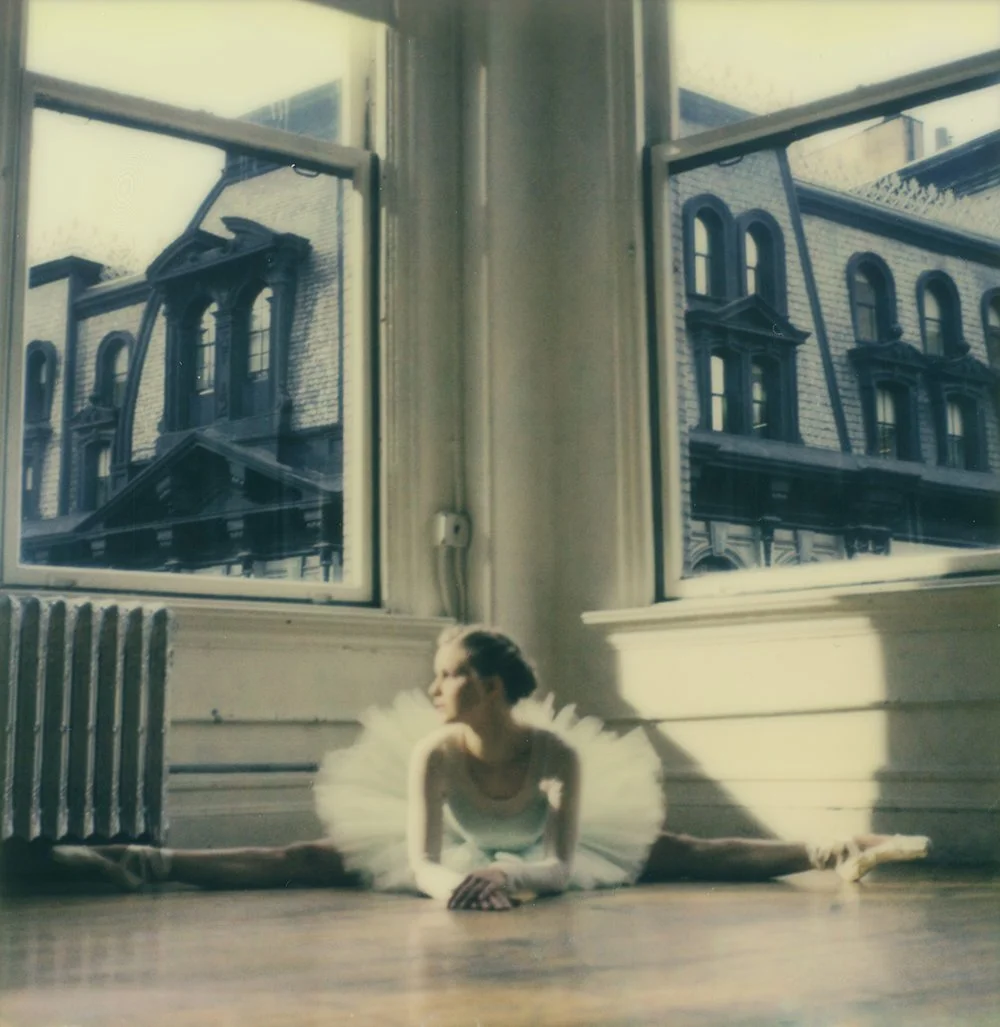 A ballet dancer in a white tutu doing a split on the floor inside a sunlit room with large windows reflecting an old building outside.