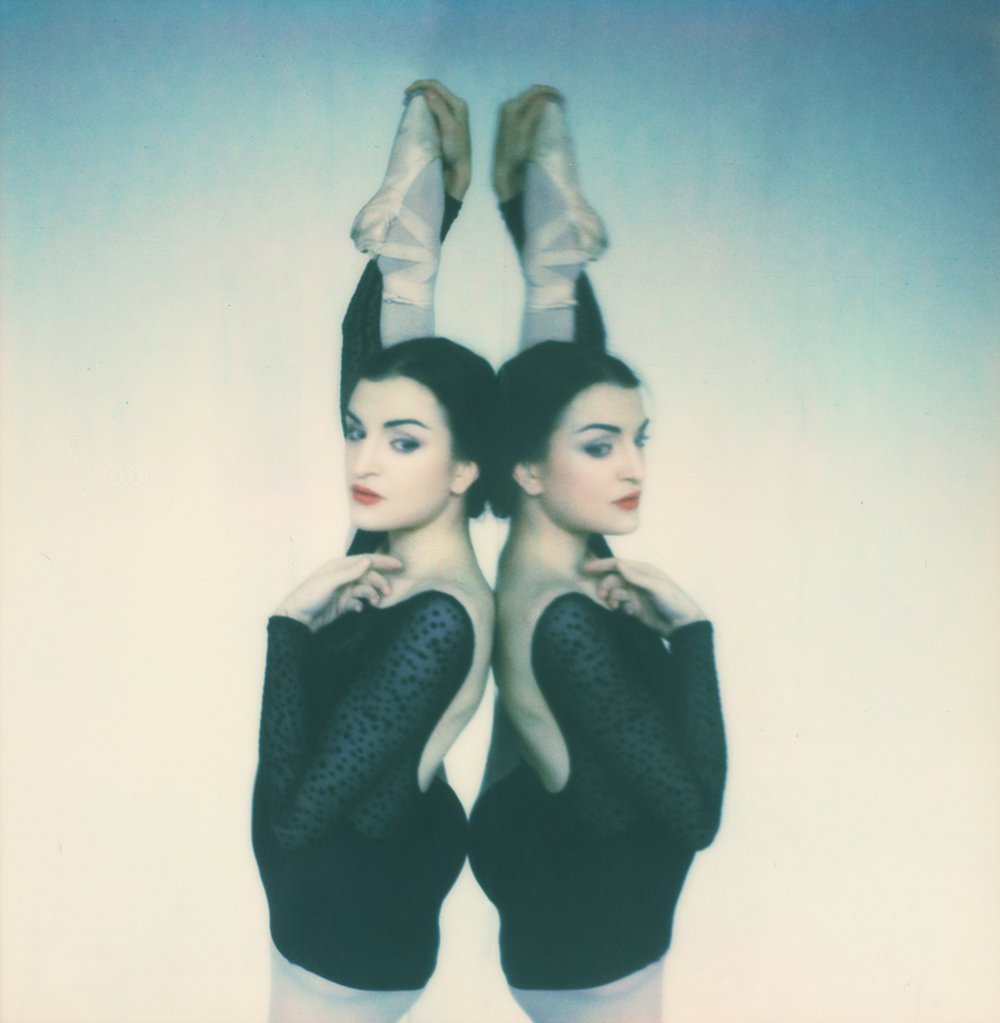 A woman in black with long sleeves, wearing white tights and ballet shoes, appears to be upside down with her head and feet touching a wall. The image is mirrored, showing two women with similar appearance, one facing forward with her hand near her c
