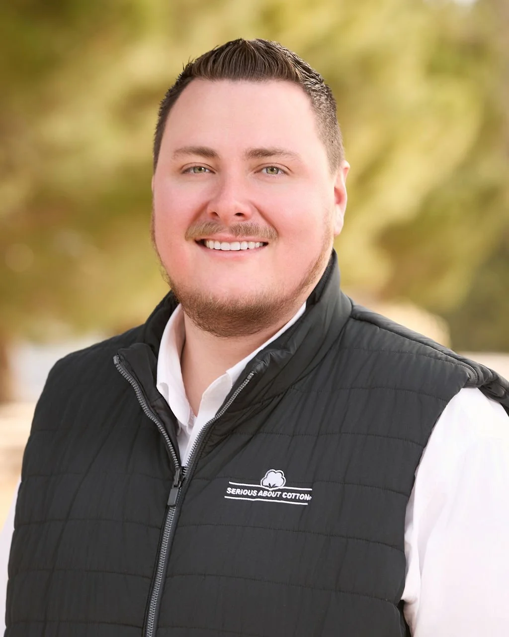 A man smiling outdoors in autumn, wearing a black vest with a logo that says 'Serious About Cotton' over a white collared shirt.