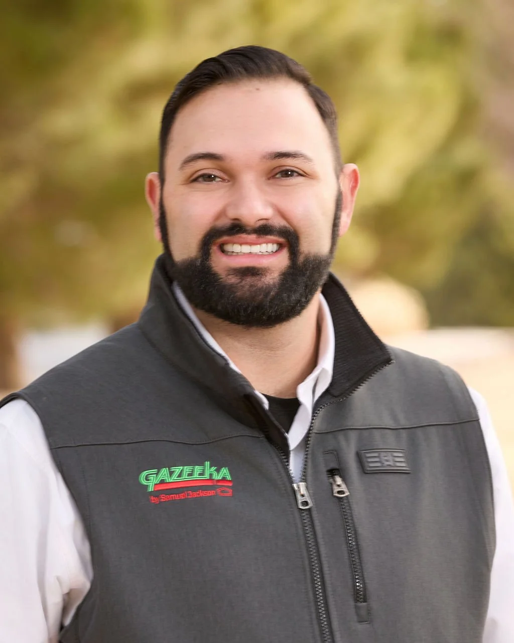 A smiling man with a beard and mustache, wearing a gray vest over a white shirt, standing outdoors with blurred trees in the background.