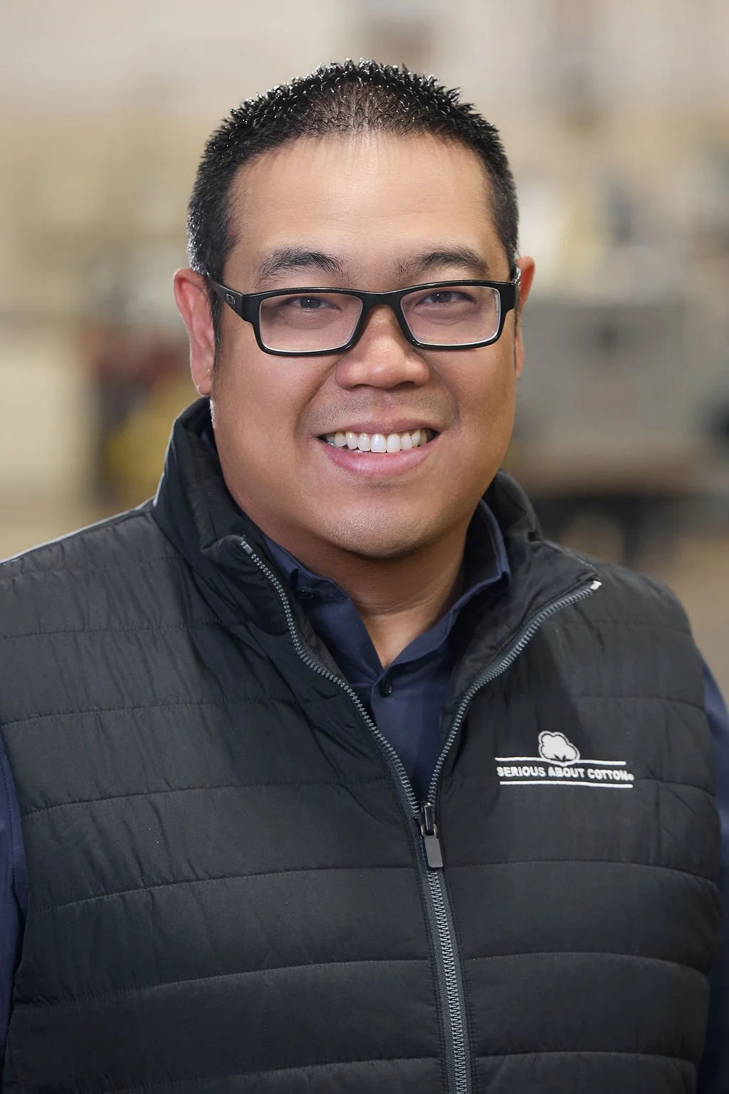 A man wearing glasses and a black puffer vest smiling at the camera, indoor setting with blurred background.