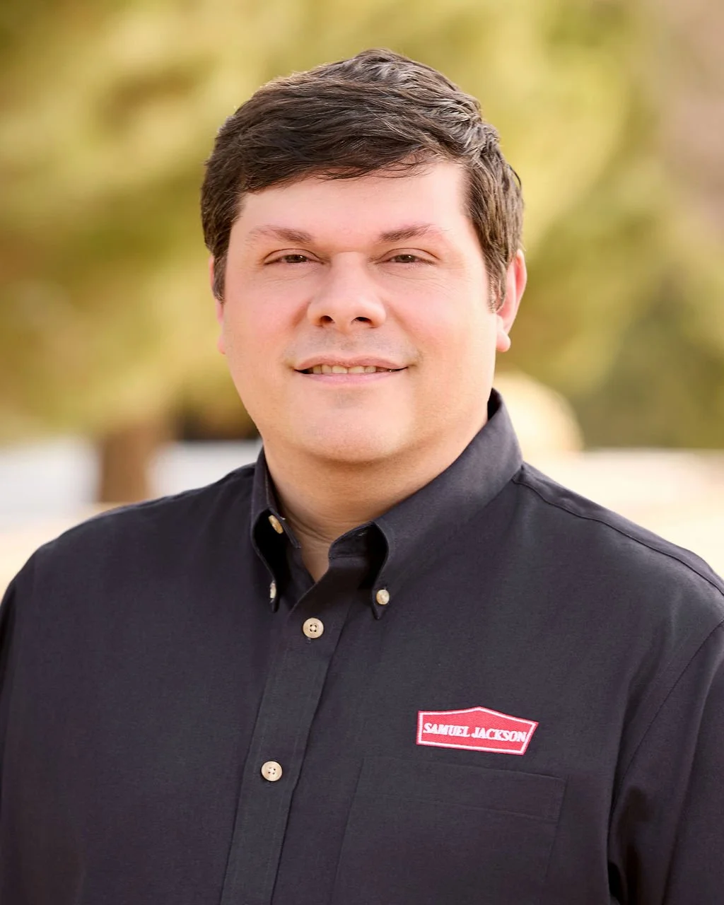 A man with short brown hair and fair skin smiling outdoors, wearing a black collared shirt with a name tag that reads Samuel Jackson, with a blurred natural background.
