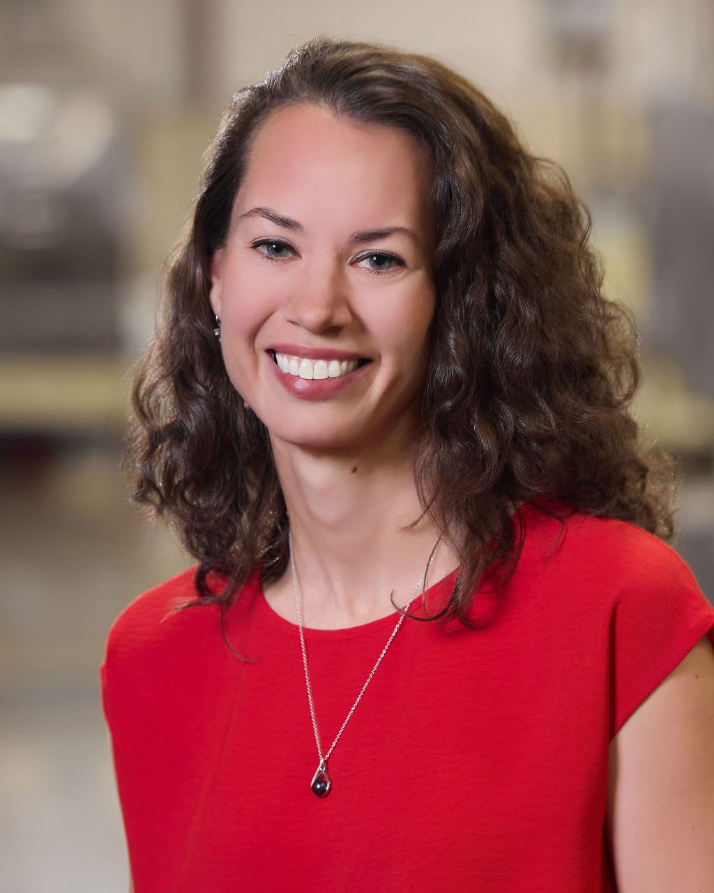 A woman with curly brown hair, wearing a red top and a silver necklace with a black pendant, smiling in an indoor setting.