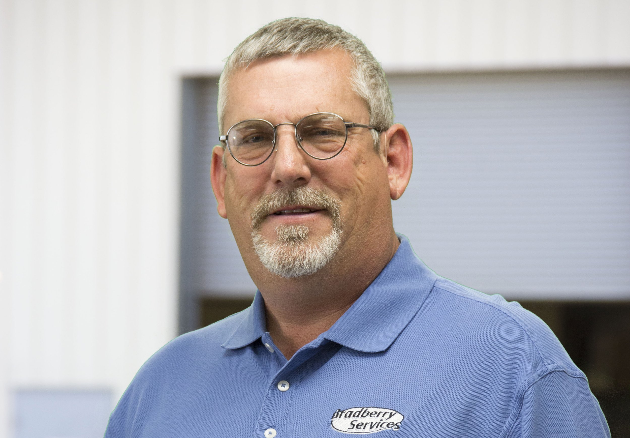 A smiling man with a bald head, blue eyes, and a full beard. He is wearing a white shirt and a black vest with logos, standing in an industrial or warehouse setting.