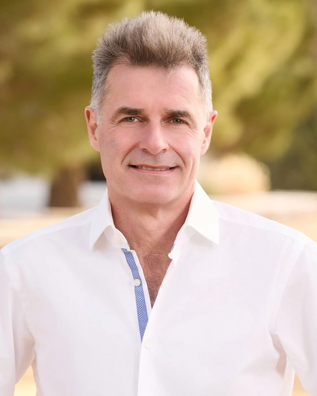 Portrait of a middle-aged man with gray hair, wearing a white button-up shirt outdoors with blurred trees in the background.