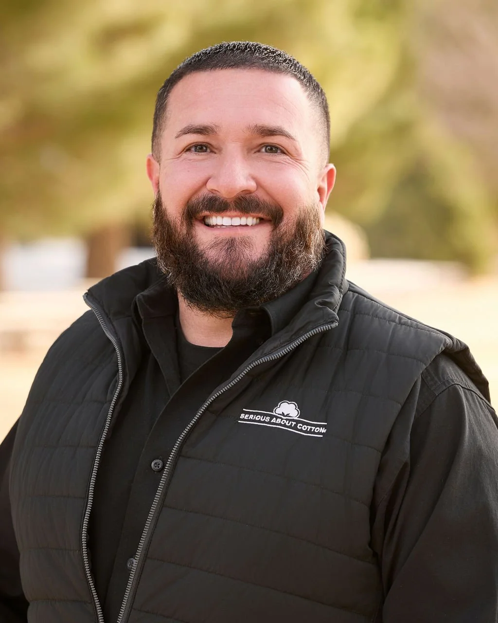 A smiling man with a beard wearing a black vest and black shirt, outdoors with a blurred background of trees.