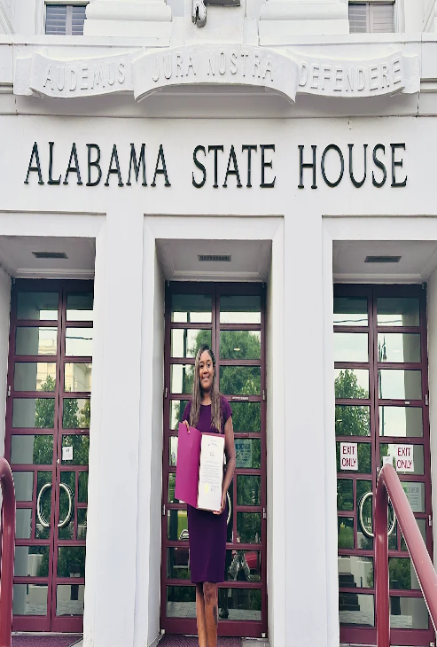 A woman is standing in front of the Alabama State House, holding a document and a pink folder, smiling at the camera.