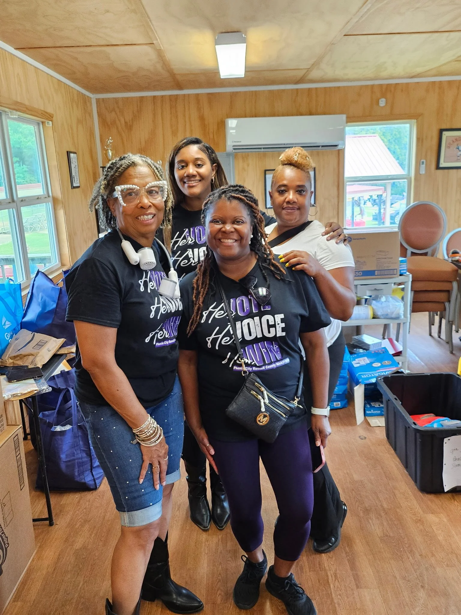 Four women standing together indoors, smiling at the camera. All are wearing black T-shirts with purple and white text, and the room has wood-paneled walls, windows, and various boxes and items in the background.