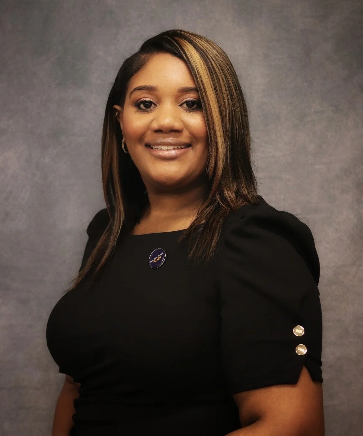 A woman with shoulder-length dark hair with blonde highlights, wearing a black dress with gold buttons on the sleeves and a NASA pin, smiling against a plain gray background.