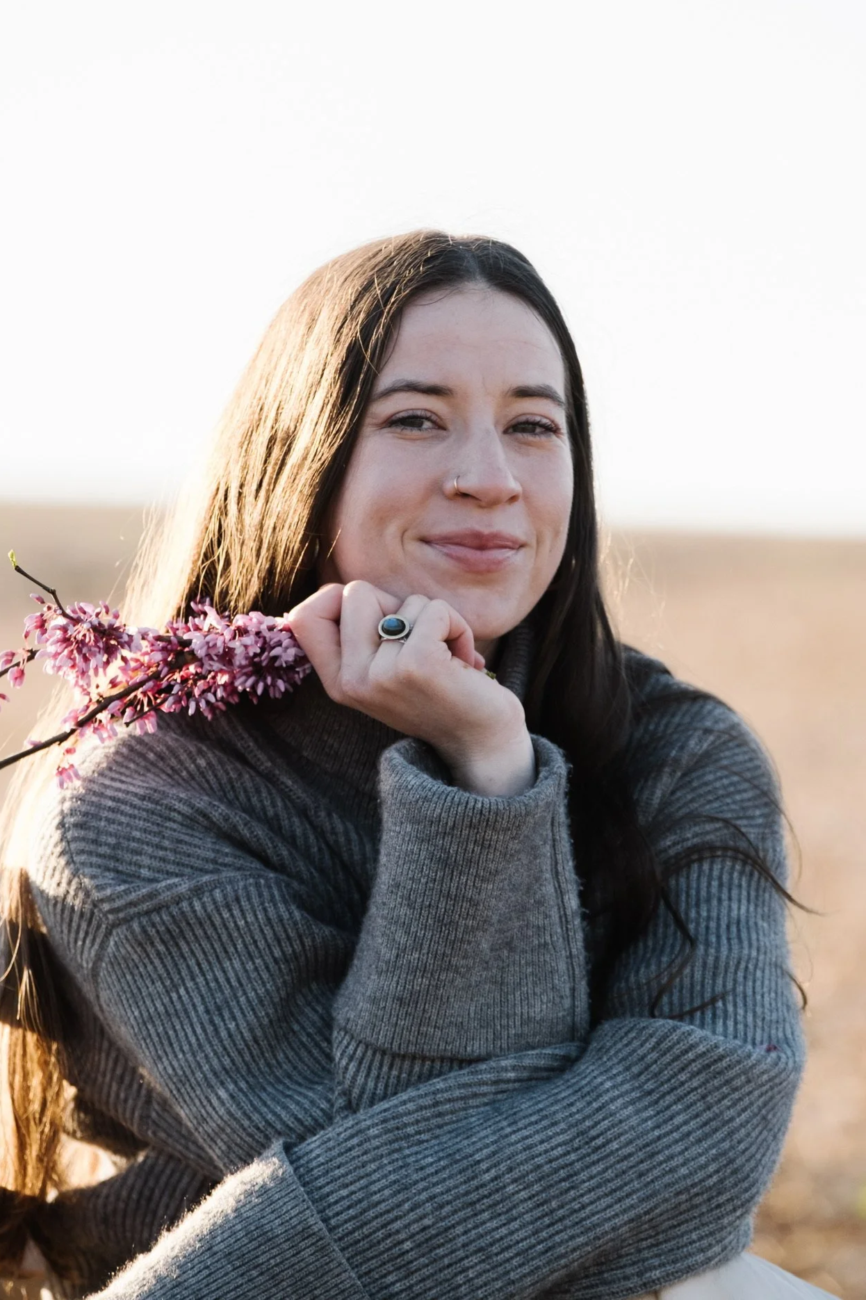 A young woman with long dark hair and light skin holding a branch of pink blossoms, smiling at the camera outdoors during sunrise.