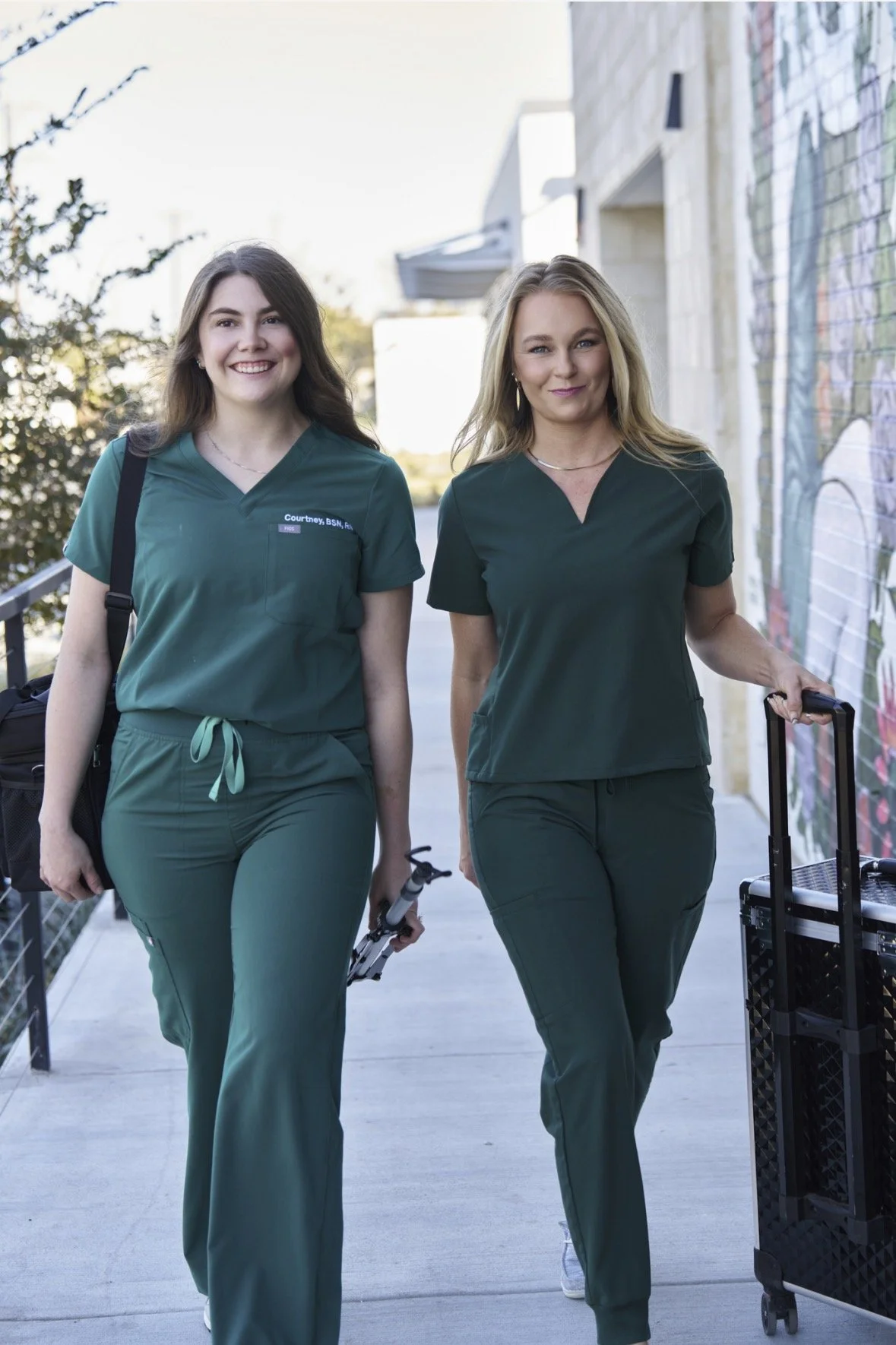 Two women in matching green medical scrubs walking outdoors, smiling, with one carrying a rolling suitcase and the other holding a cane and a drink tray.