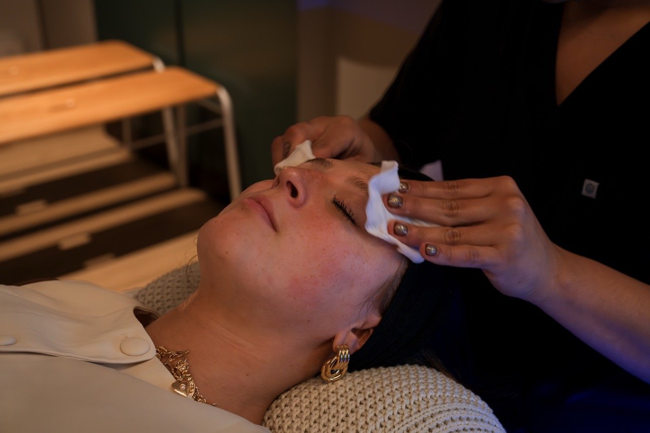 A woman receiving a facial treatment, lying down with her eyes closed, as a professional gently wipes her face with a cloth.