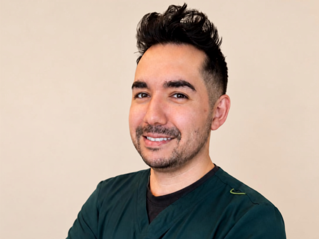 A man with dark, styled hair and a goatee, wearing scrubs, smiling against a plain beige background.