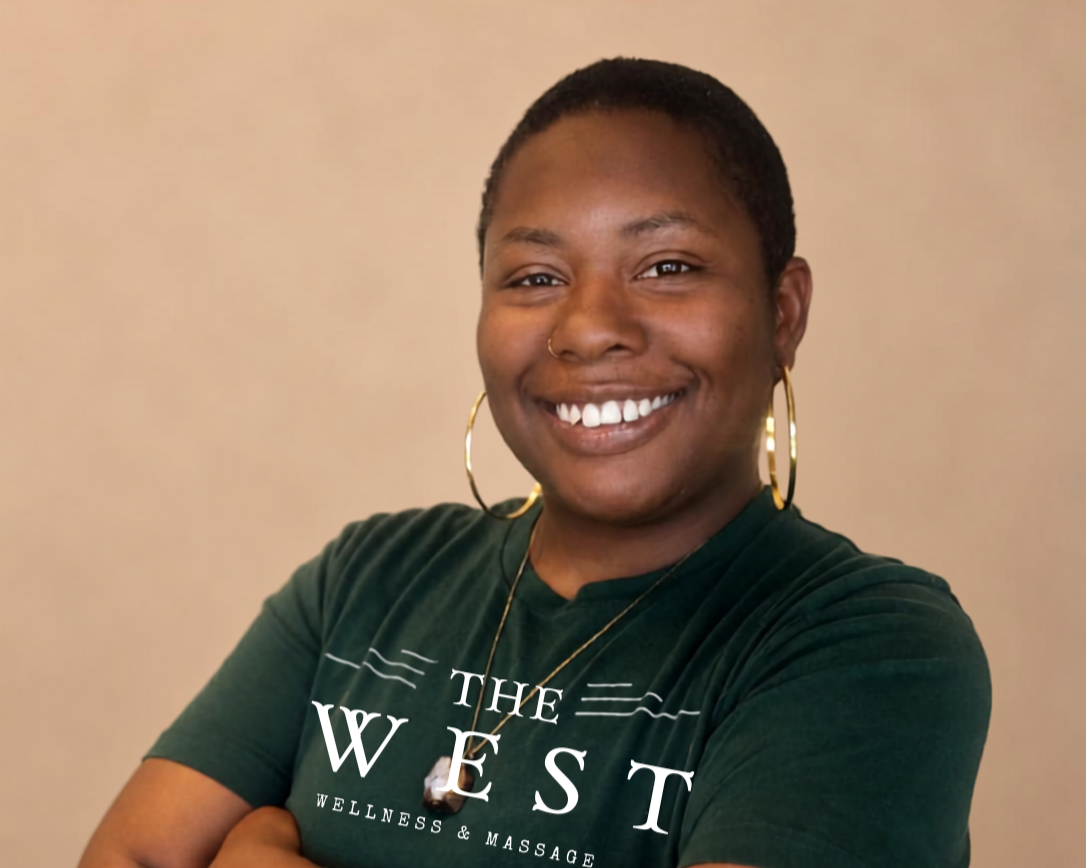 A smiling woman with short hair, wearing gold hoop earrings, a nose ring, and a dark green T-shirt with the words "The West Wellness & Massage" printed on it, standing against a neutral background.