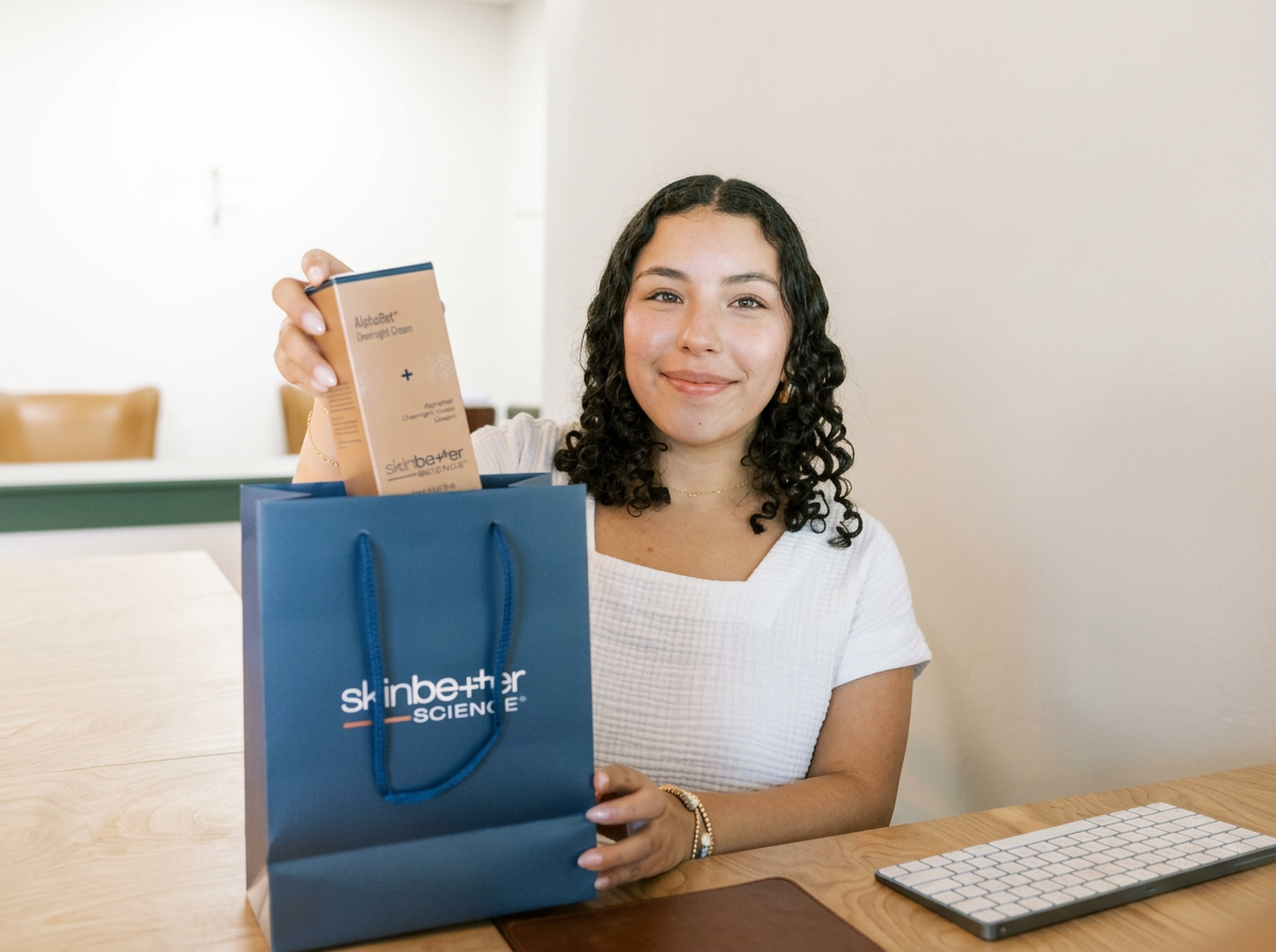 A woman with curly dark hair smiling at the camera, holding a skincare product box and a blue gift bag with 'skin better SCIENCE' written on it, sitting at a wooden desk with a keyboard in front of her.