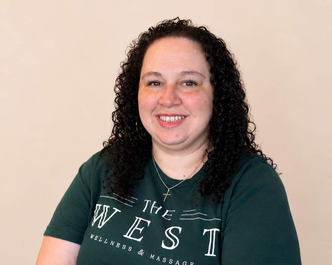 A woman with curly dark hair smiling, wearing a green T-shirt that says "The West Wellness & Massage," and a silver cross necklace, standing against a plain light-colored background.