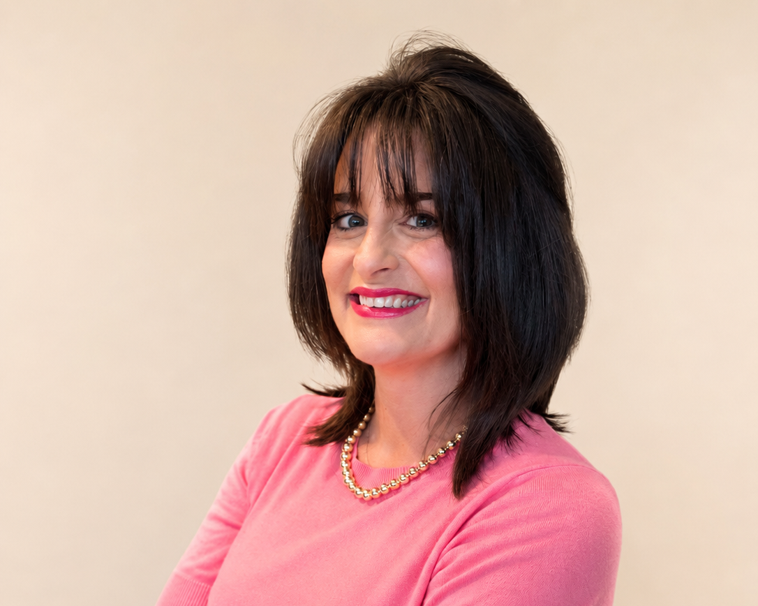 A woman with dark brown hair and bangs, wearing a pink top and a pearl necklace, smiling against a plain background.