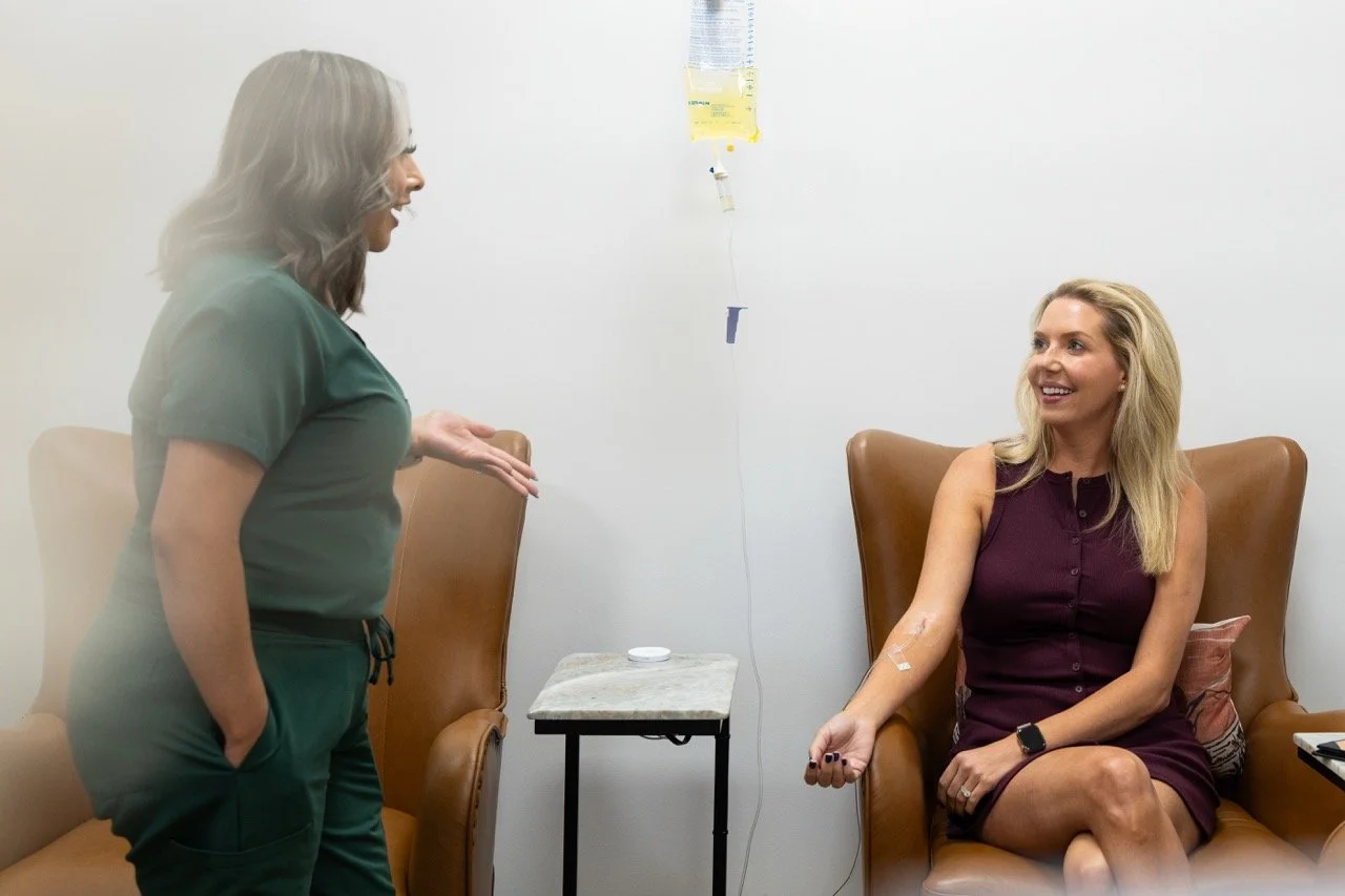 A woman in hospital scrubs talking to a woman sitting in a brown chair with an IV inserted in her arm in a medical room.