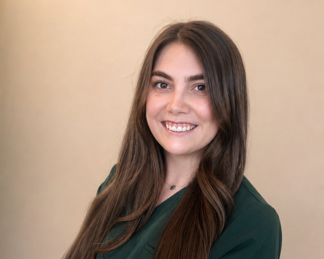 Portrait of a young woman with long brown hair, smiling, wearing a green top, standing against a beige background.