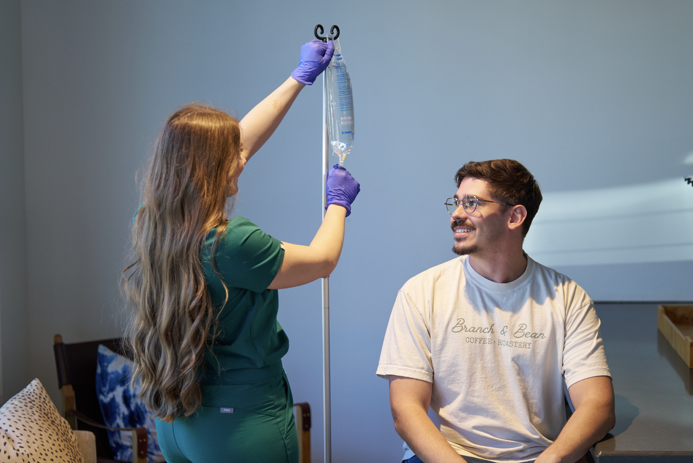 Nurse hanging an IV bag during an at-home IV therapy visit for a smiling client from The West Wellness and Massage in Fort Worth
