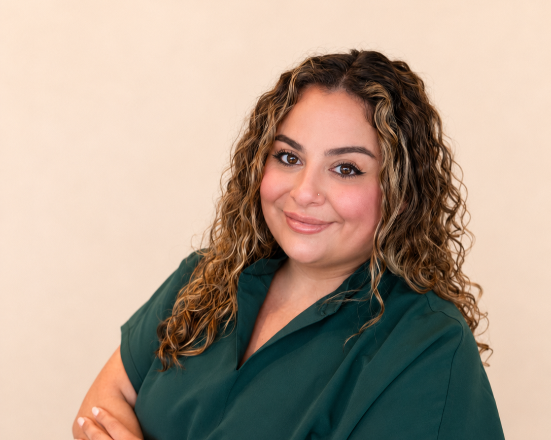 A woman with curly hair and a nose piercing, wearing a dark green top, smiling with arms crossed against a plain beige background.