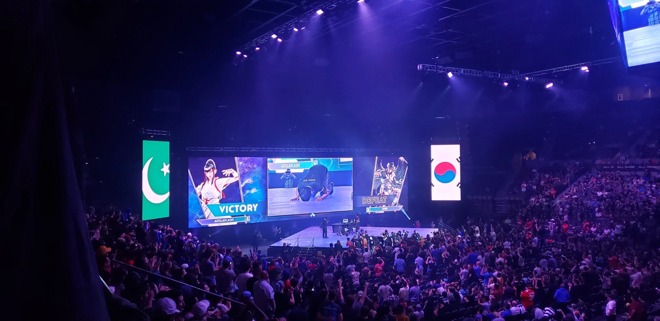 An indoor sports arena filled with spectators watching a video game competition on a large screen. The screen displays a player named Arslan Ash with the words 'Victory' and 'Defeat' shown.