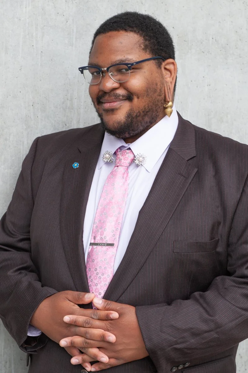 A man wearing a dark suit, pink tie, and glasses, standing with hands clasped in front of a gray wall, smiling at the camera. He has earrings and decorative pins on his shirt.