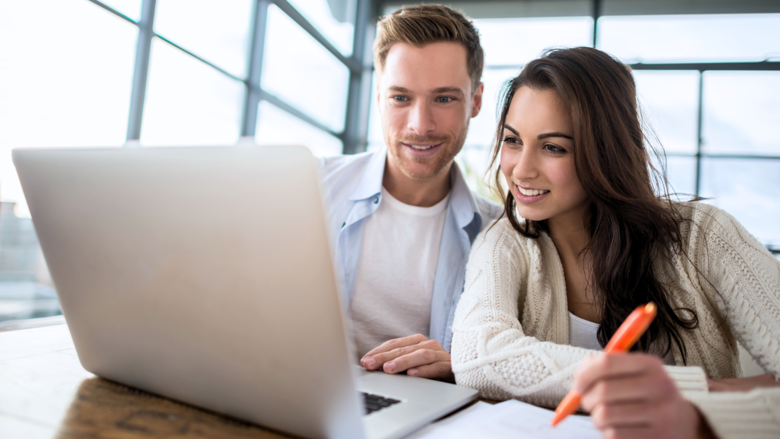 Two young adults, a man and a woman, smiling and looking at a laptop screen together in a bright, modern office space.