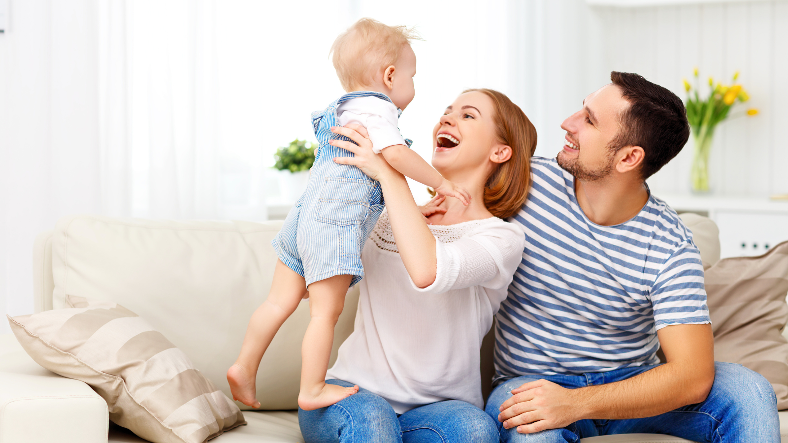 A family of three, a woman, a man, and a young child, sitting on a cream-colored couch in a bright room. The woman is holding the child, who is lifting his hands toward her, and all are smiling joyfully.