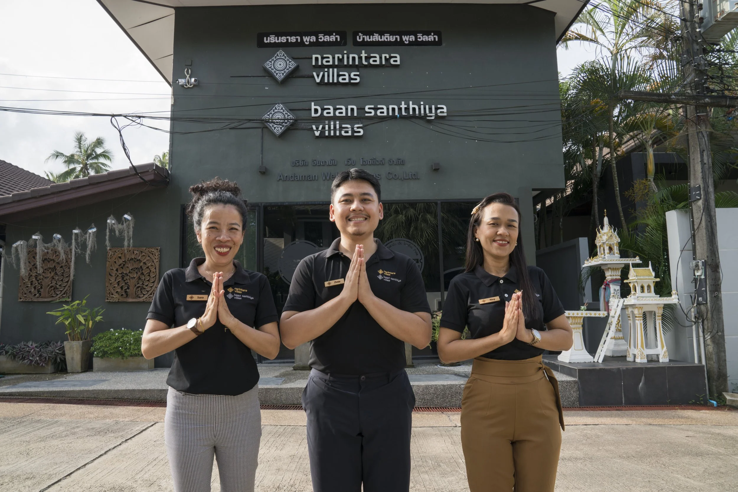 Three hotel staff members stand outside Narintara Villas and Baan Santhiya Villas, smiling and holding their hands together in a traditional greeting. The background includes the hotel building, decorative elements, plants, and a small shrine.