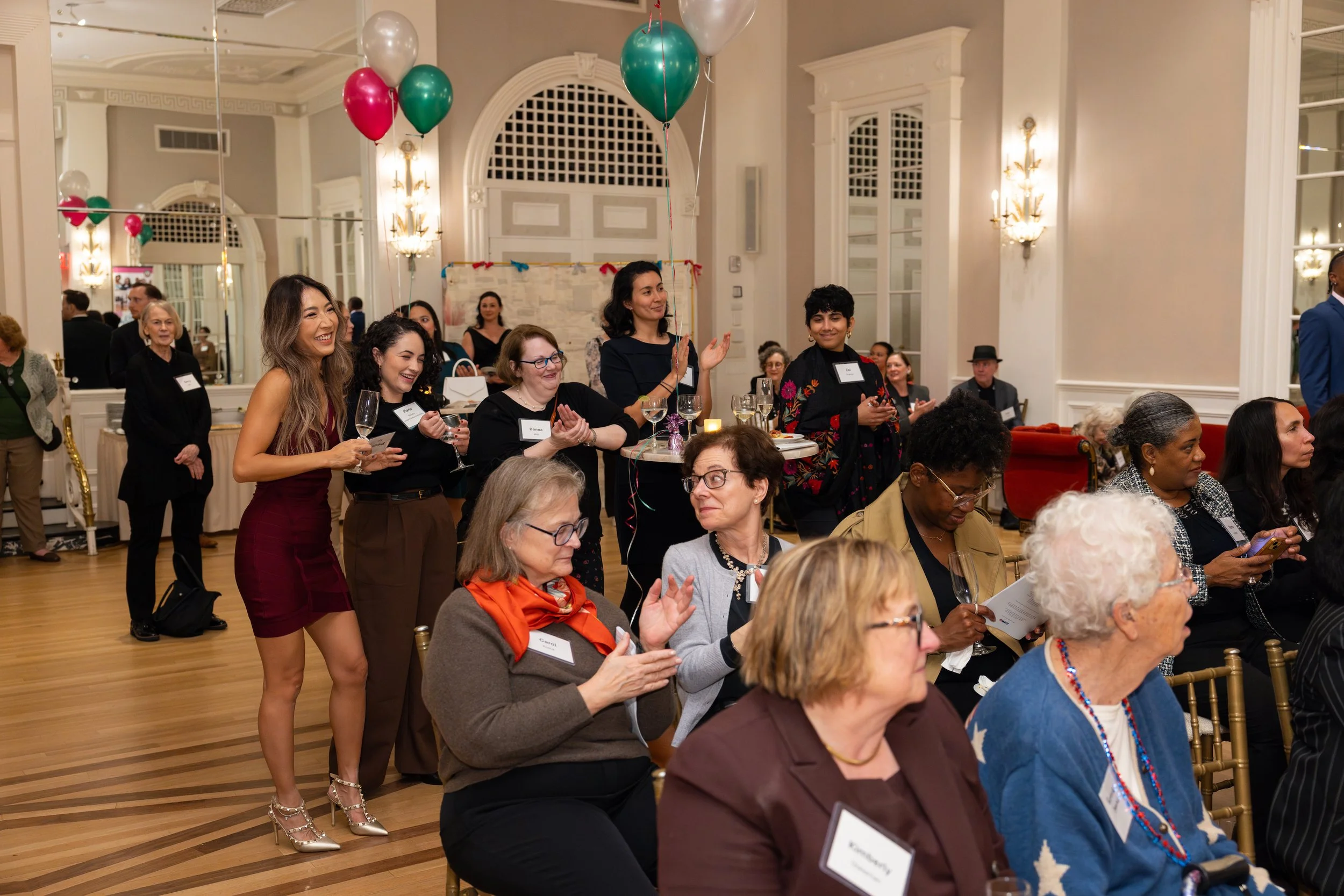 Group of women in an elegant formal event, some are clapping and smiling, with balloons and refreshments on tables, in a decorated banquet hall.