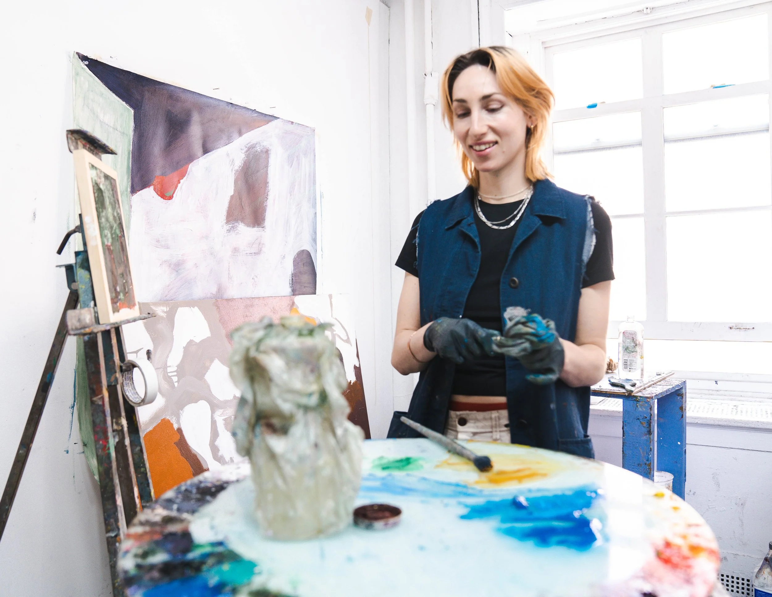 A woman in a blue vest and black shirt paints in an art studio, standing in front of a large abstract painting on the wall.